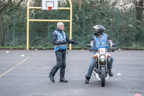 An instructor gestures while talking to a learner sitting on a white motorbike in front of a yellow basketball hoop.
