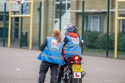 An instructor in a blue vest, seen from behind, speaks to a learner on a red motorbike with a yellow number plate.