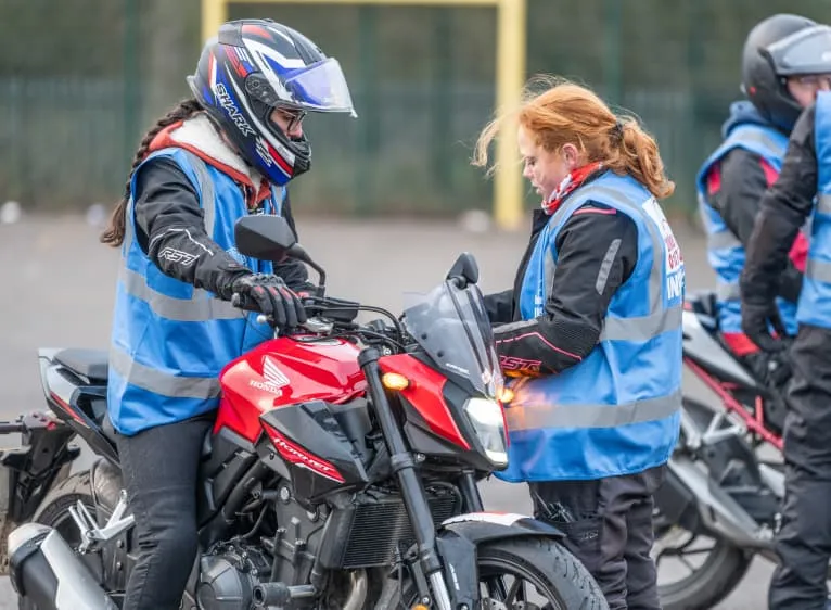 An instructor in a blue vest checks the controls of a red motorbike while a learner sits on it.