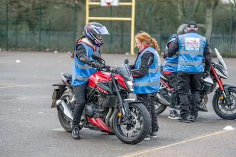 Learners and instructors gather around a red motorbike while a learner in a blue-and-red helmet sits on it.