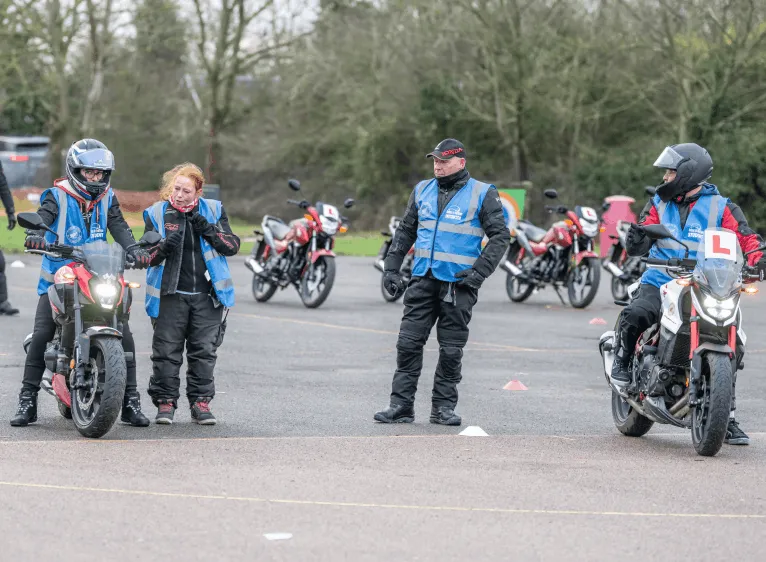 An instructor talks to a learner while two others wait on their bikes, with parked red bikes behind them.
