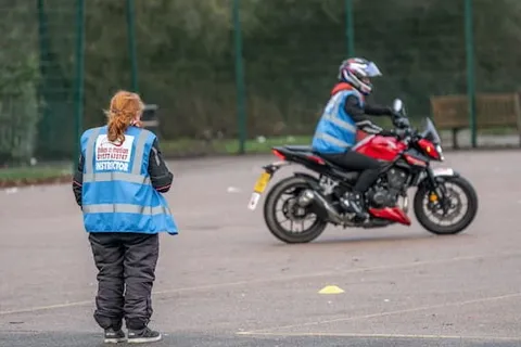 An instructor stands on the practice area as a learner rides a red motorbike in the distance.