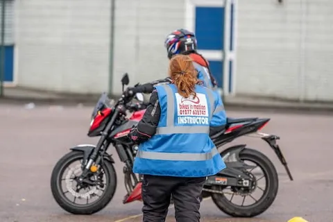 An instructor in a blue vest, seen from behind, faces a learner on a red motorbike in front of a white building.