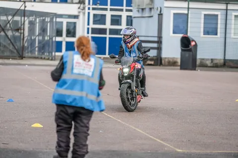 An instructor in a blue vest, seen from behind, watches a learner approach on a dark motorbike with its headlight on.