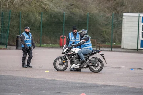 A white motorbike carrying two people rides past an instructor in a blue vest standing on the practice area.