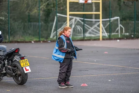An instructor in a blue vest stands beside a parked motorbike with a yellow number plate, with goalposts behind.