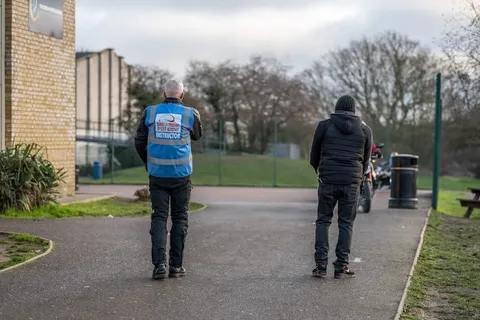 An instructor in a blue vest and another person stand with their backs to the camera beside a brick building.