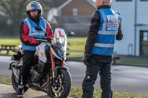 An instructor in a blue vest talks to a learner on a white and red motorbike, with houses and a green field behind.