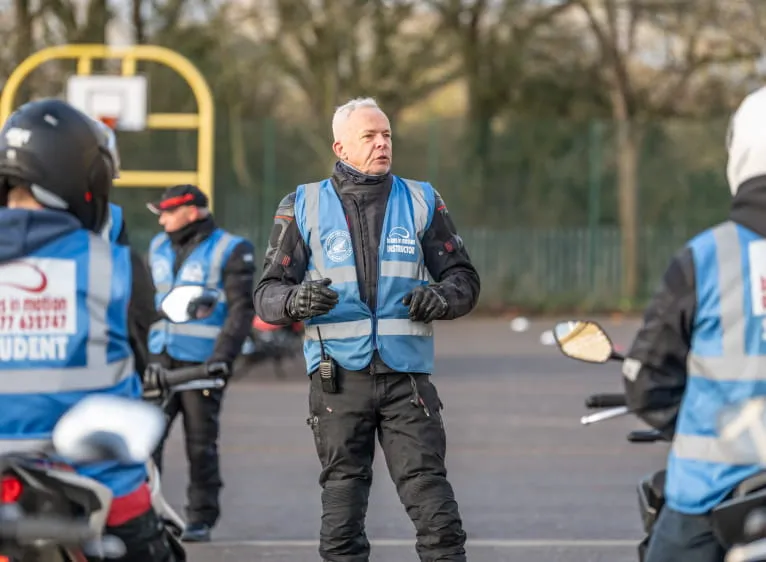 An instructor in a blue Bikes in Motion vest talks to learners on their bikes, who are seen from behind in blue student vests.