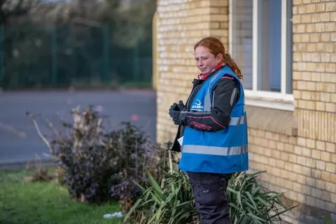 A smiling instructor with red hair stands in a blue vest against a yellow-brick wall, beside a green shrub.