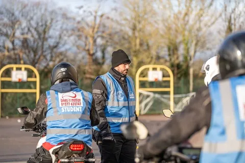A learner on a bike and an instructor in a beanie talk beside a yellow basketball hoop, with another rider in the foreground.