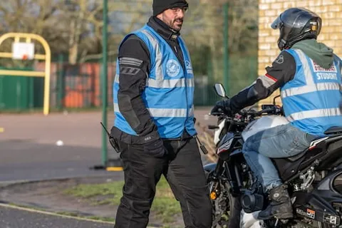 An instructor in a beanie and blue vest walks towards a learner on a bike in the sunshine.