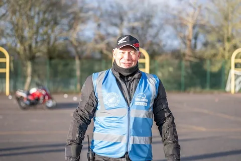 A smiling instructor in a blue vest and a black cap stands in front of the training ground, with a red bike parked behind.