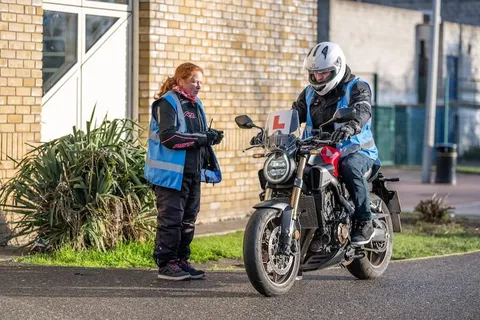 An instructor with red hair stands beside a learner sitting on a dark motorbike in front of a yellow-brick building.