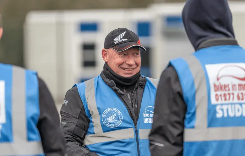 A smiling instructor in a black Honda cap and blue vest stands between two learners who are seen from behind in student vests.