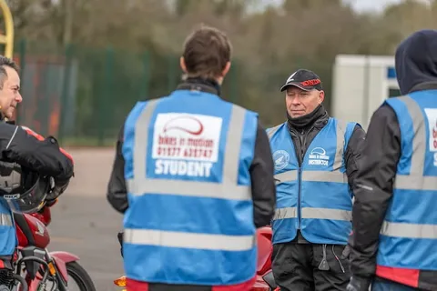Learners in blue student vests, seen from behind, listen to an instructor in a black cap at a briefing.