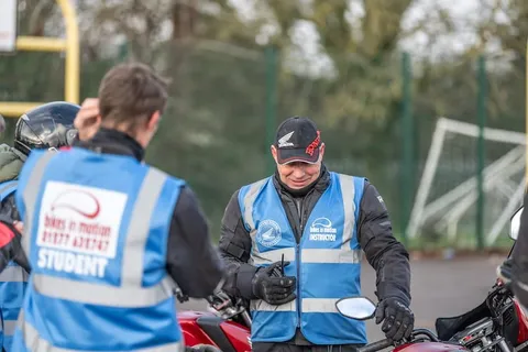 A learner in a student vest, seen from behind, talks with an instructor in a black cap beside a red bike.