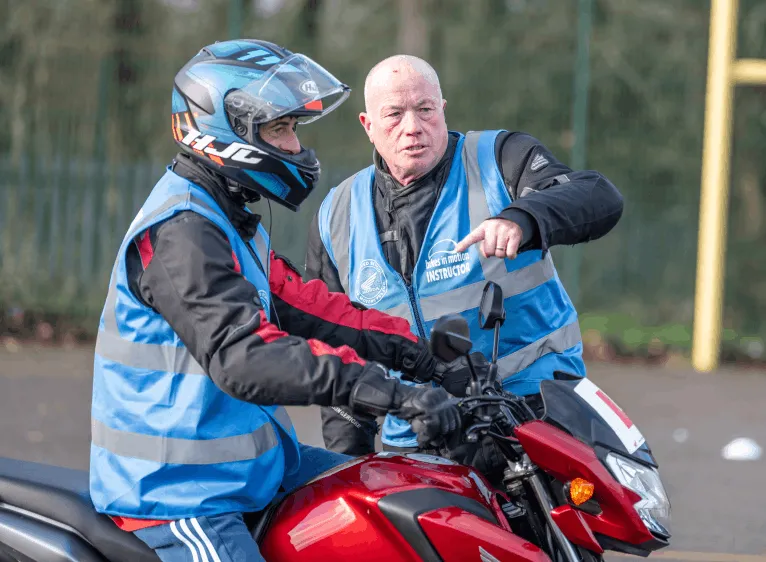 An instructor in a blue vest points ahead while a learner in a blue helmet sits on a red motorbike.