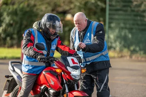 An instructor points out a feature on a red motorbike to a learner in a full-face helmet.