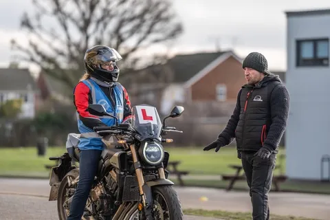 A learner in a full-face helmet sits on a dark motorbike talking to an instructor in a beanie who stands beside it.