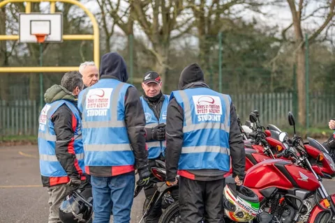Learners and instructors in blue vests gather around a line of red motorbikes.