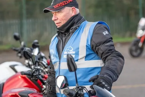An instructor in a black cap looks down at the controls of a red motorbike.