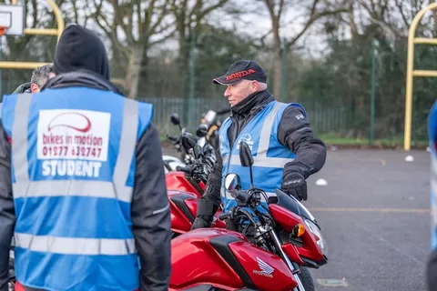 An instructor in a black cap stands beside a red motorbike while a learner in a student vest looks on.