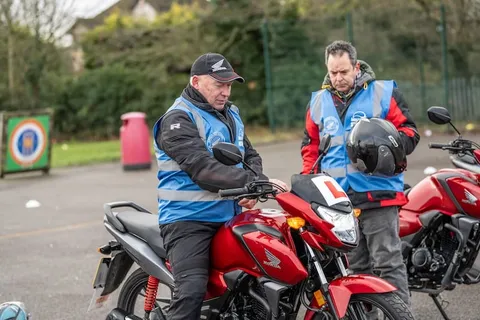 An instructor in a cap explains the controls to a learner sitting on a red motorbike while another instructor watches.