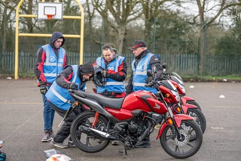 A group of learners and instructors crouch and stand around a red motorbike, checking it over.
