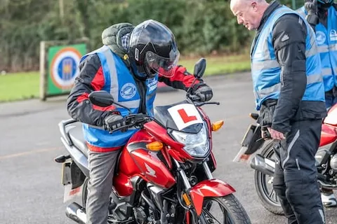 An instructor checks a learner sitting on a red motorbike with an L plate, while another rider waits behind.