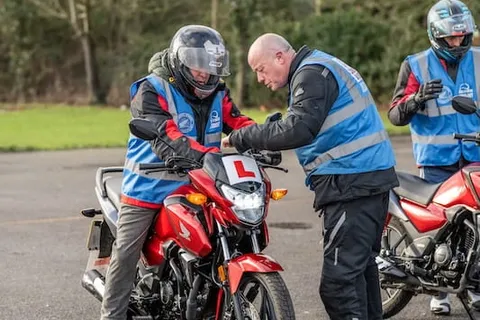 An instructor leans in to check a learner's red motorbike while another rider waits behind them.