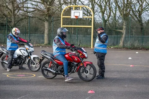 A learner on a red motorbike waits beside an instructor in a cap, while another learner sits on a white bike.
