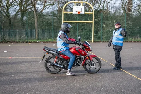 A learner on a red motorbike pauses near an instructor in a cap who stands to the right.