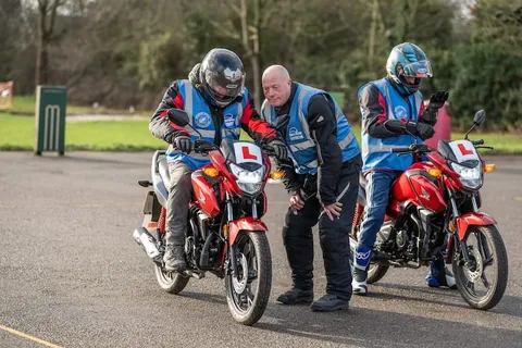 An instructor leans in to help a learner on a red motorbike while a second learner on another red bike looks on.