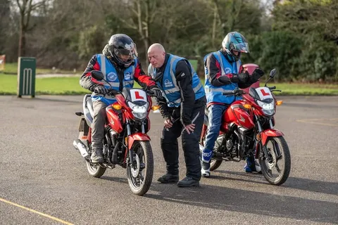An instructor bends to talk with a learner on a red motorbike while another learner looks on.