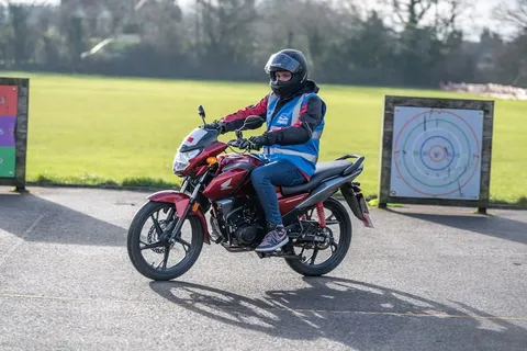 A learner in a black helmet rides a red motorbike across sunlit tarmac, with a target board behind.