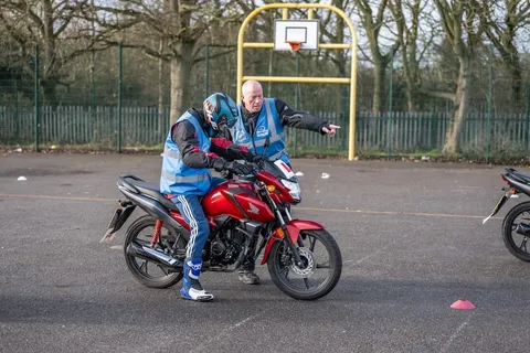 A learner sits on a red motorbike while an instructor behind them points across the practice area.