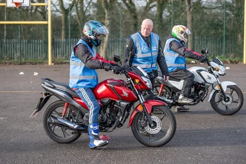 A learner on a red motorbike waits with an instructor and another learner on a white bike behind.