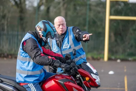 An instructor in a blue vest points ahead while a learner in a teal helmet sits on a red motorbike.