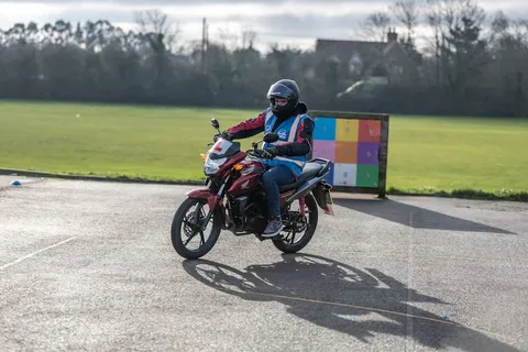 A learner in a black helmet rides a red motorbike across a wide open area, with a colourful target board behind.