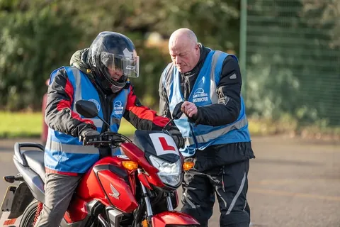 An instructor leans over a red motorbike to point something out to a learner in a helmet with a visor.