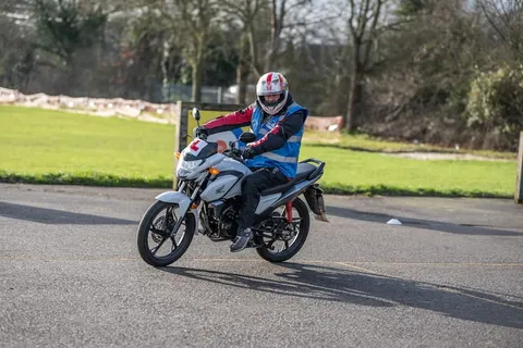 A learner in a white helmet rides a silver motorbike across the practice area, with a green field behind.
