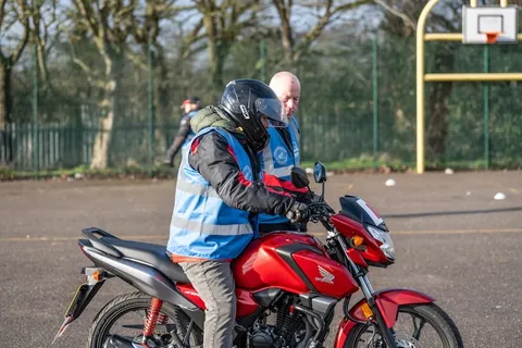 A learner in a black helmet sits on a red motorbike with an instructor standing just behind them.