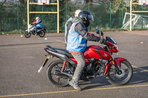 A learner in a black helmet sits on a red motorbike, with another rider on a white bike in the distance.