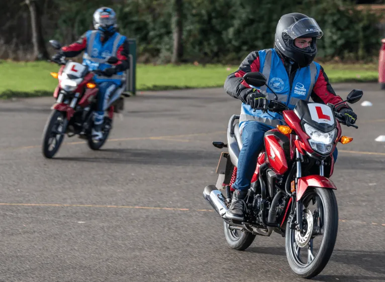 A learner in a blue student vest and black helmet rides a red motorbike towards the camera, with a second learner following behind.