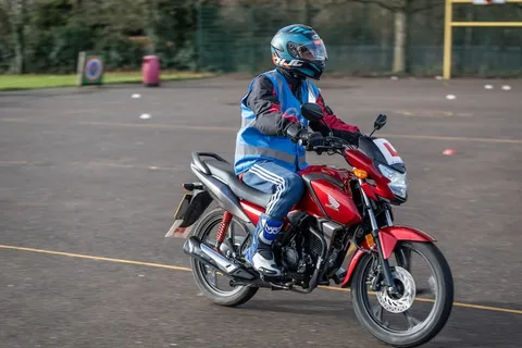 A learner in a teal helmet and blue vest rides a red motorbike with an L plate towards the camera.