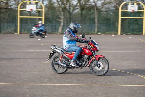 A learner in a black helmet rides a red motorbike across yellow court markings, with another bike in the distance.