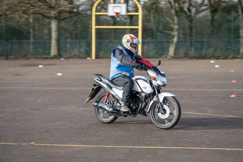 A learner in a colourful helmet rides a white motorbike across the yellow-marked practice area.