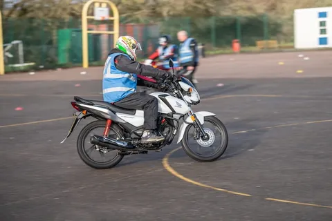 A learner in a green and white helmet turns a white motorbike in front of basketball hoops, with other riders behind.