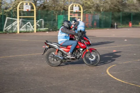 A learner in a black helmet leans a red motorbike into a turn, with basketball hoops and goalposts behind.